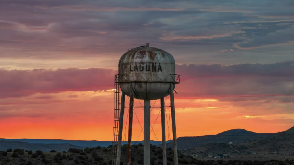 Rusty water tower labeled 'LAGUNA' stands tall against a vibrant sunset sky with scattered clouds over a desert landscape.