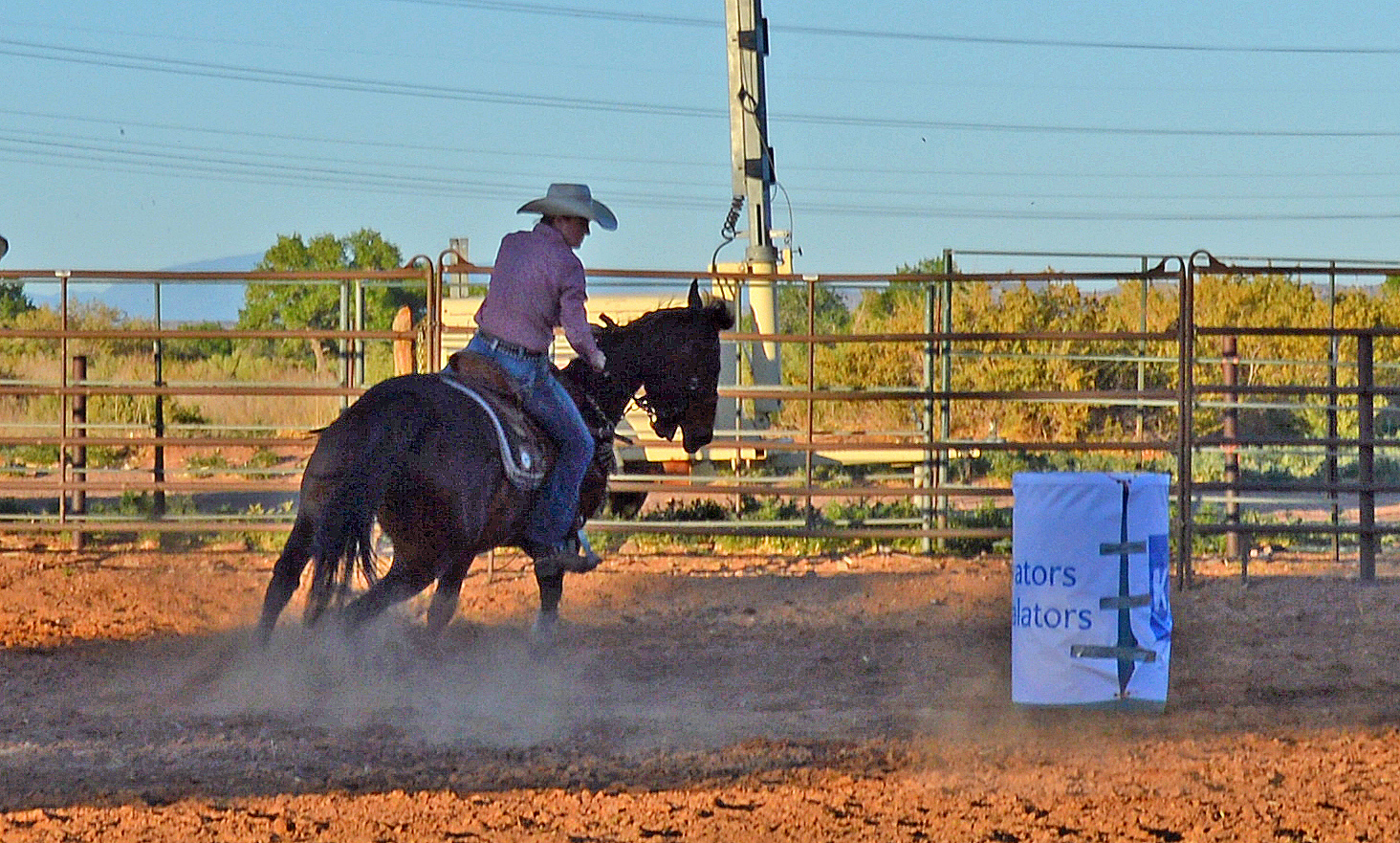 Cowboy in a hat riding a dark horse swiftly around a barrel in a dusty rodeo arena.