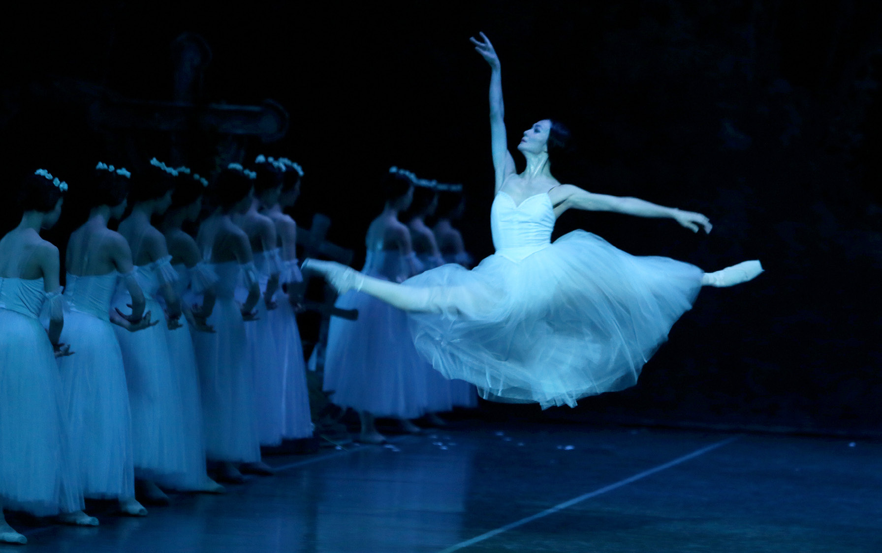 Ballerina in a white tutu performing a grand jeté leap on stage with dancers lined up in the background.