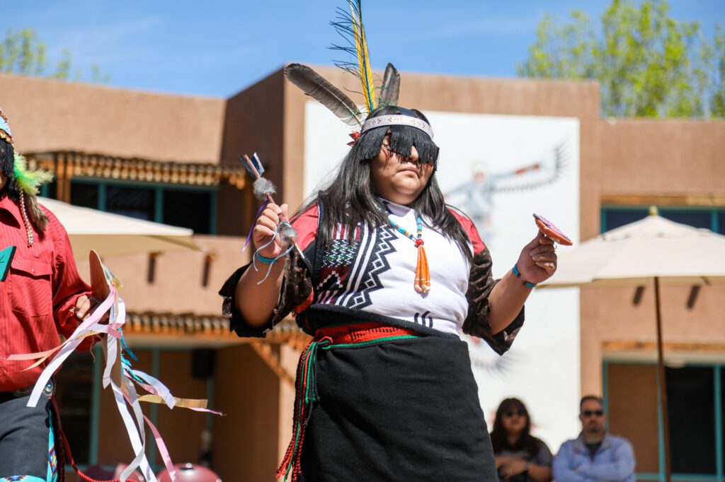 Native American woman in traditional attire performs a ceremonial dance outdoors with feathers and rattles.