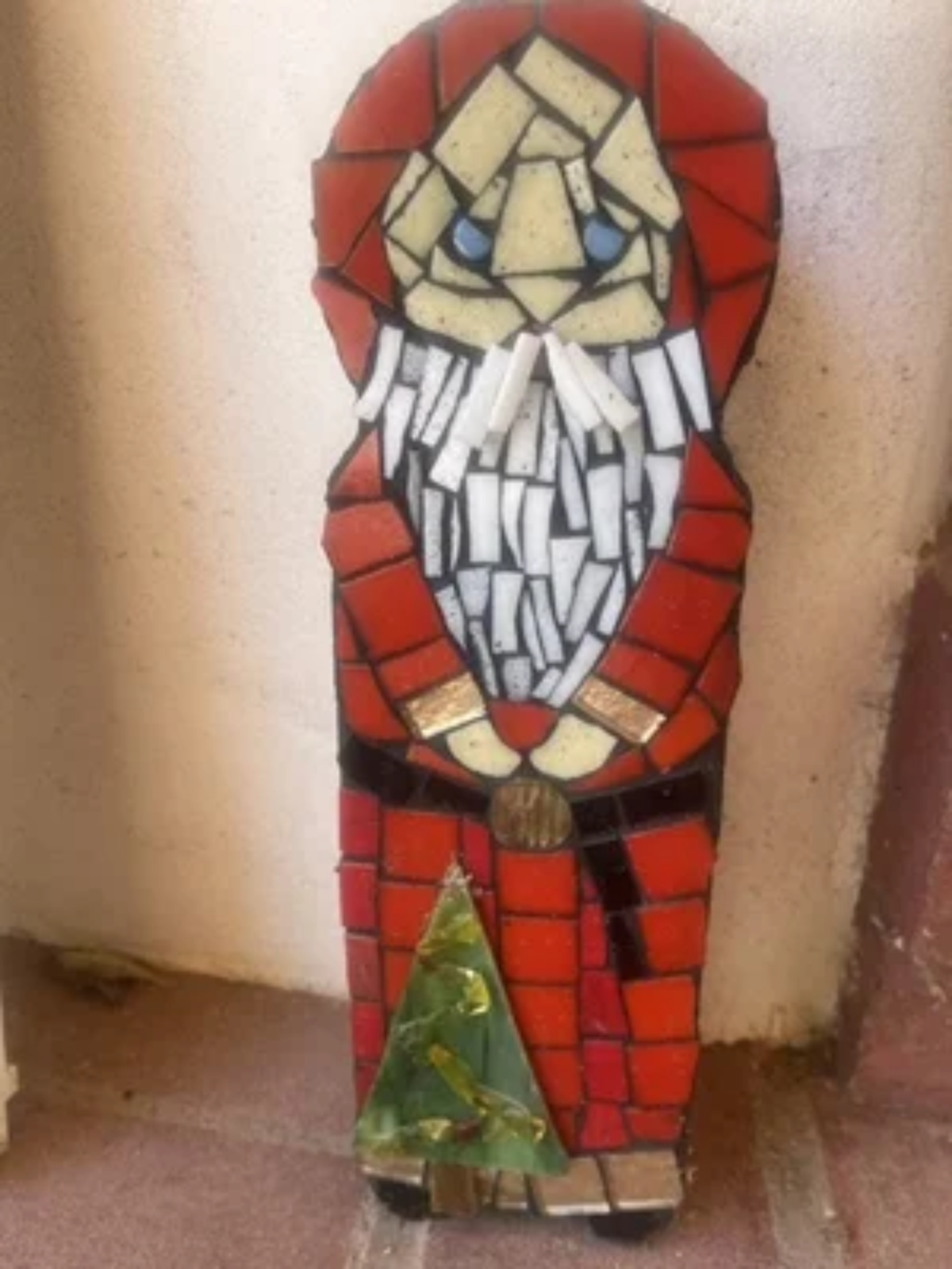Mosaic artwork of Santa Claus in red suit holding a green Christmas tree, made with colorful tiles on a wall corner.