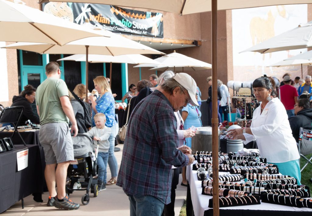 Shoppers browse handcrafted jewelry displayed on tables under umbrellas at an outdoor market with diverse visitors in casual attire.