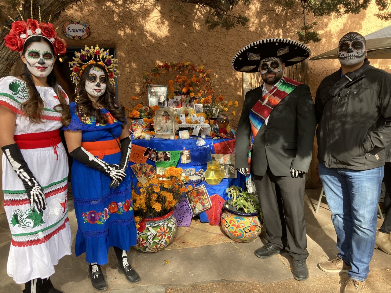 Four people in Día de los Muertos attire stand beside a colorful outdoor altar decorated with flowers, candles, and offerings.
