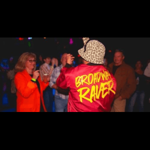 A person in a leopard-print hat and red jacket labeled “BROWN RAVER” dances among a crowd in a dimly lit room.